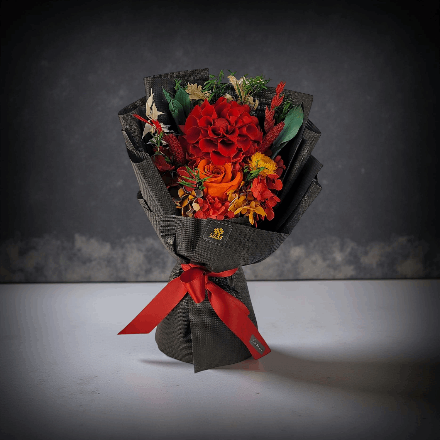 Luxurious preserved flower bouquet in black wrap with red and orange blooms, including dahlia, rose, and hydrangea, tied with red ribbon – Luxe Flowers